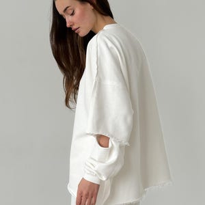 Puede incluir: Una sudadera blanca de gran tamaño con bordes sin rematar en las mangas y el dobladillo. Las mangas largas tienen un detalle de corte único. El conjunto forma parte de un conjunto a juego, lo que sugiere un estilo casual y cómodo. La tela parece ser un material suave y de peso medio.