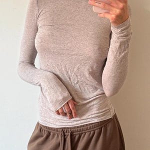 以下が含まれることがあります： ボートネックのライトグレーの長袖シャツ。シャツは茶色のスウェットパンツと組み合わせています。生地は柔らかく、わずかにテクスチャード加工されているように見えます。全体的なスタイルはカジュアルで快適です。
