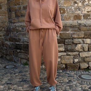 Könnte beinhalten: Ein altrosa Set bestehend aus einem langärmeligen Kapuzenpullover mit Kängurutasche auf der Vorderseite und einer passenden Jogginghose mit Kordelzug. Der Hoodie hat eine Kapuze mit Kordelzug und die Hose eine lockere Passform mit Kordelzug.