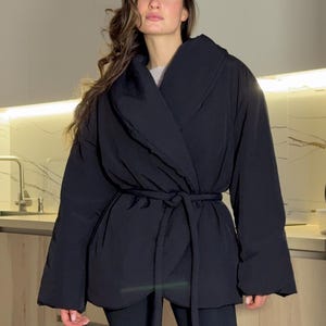 Puede incluir: Chaqueta envolvente negra con cuello ancho y cinturón a juego. La chaqueta tiene mangas largas y un corte relajado. La modelo lleva una camiseta gris debajo. El fondo es un entorno de cocina neutro.