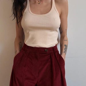 Könnte beinhalten: Ein cremefarbenes geripptes Tanktop kombiniert mit einer hoch taillierten Cordhose mit weitem Bein in einem tiefen Burgunderrot. Die Hose hat einen Knopfverschluss und Taschen. Das Outfit ist schlicht und stilvoll.