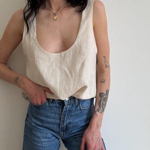 Può includere: Un top in lino color crema con scollatura rotonda, abbinato a jeans blu. È visibile una collana d'argento con un ciondolo circolare. La modella ha tatuaggi sulle braccia.