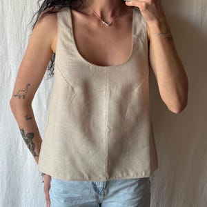 May include: A beige linen tank top with a scoop neckline. The top has a tailored fit and a slightly flared hem. The fabric appears to have a subtle texture. The model is wearing light blue jeans.