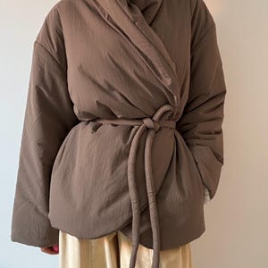 May include: A brown wrap jacket with a wide collar and a matching tie belt. The jacket has a quilted appearance and long sleeves. The model is wearing beige corduroy pants. The jacket is a neutral color and appears to be made of a soft material.