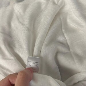 May include: A white, lightweight, semi-sheer fabric with a visible woven texture. A small white care label is attached to the fabric, featuring the text 100% tencel, along with standard laundry care symbols and the word Ukraine printed on it.
