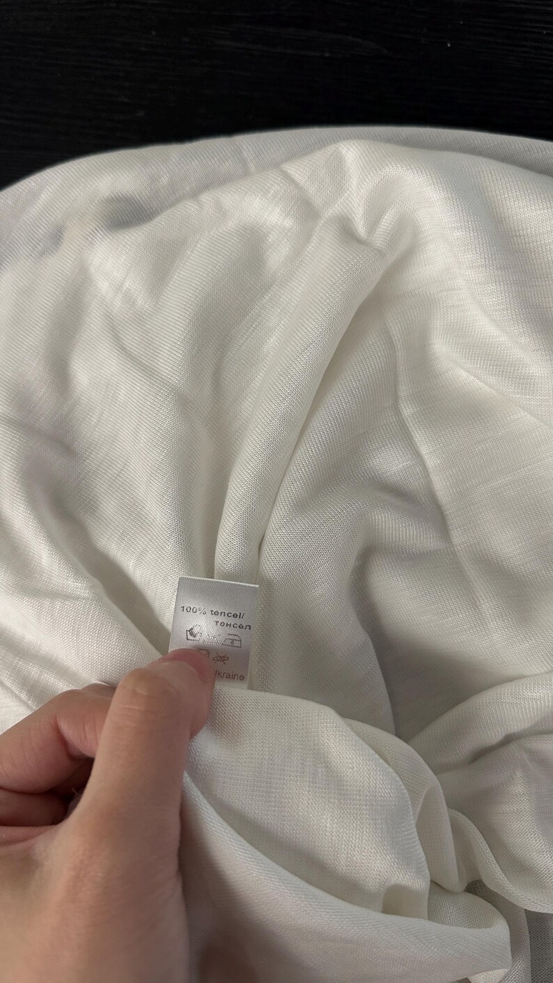 May include: A white, lightweight, semi-sheer fabric with a visible woven texture. A small white care label is attached to the fabric, featuring the text 100% tencel, along with standard laundry care symbols and the word Ukraine printed on it.