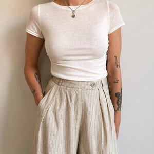 May include: A white short-sleeved crew neck t-shirt paired with high-waisted beige trousers featuring vertical pinstripes, a single button closure at the waistband, and deep front pleats. The trousers have a wide-leg silhouette and side pockets.