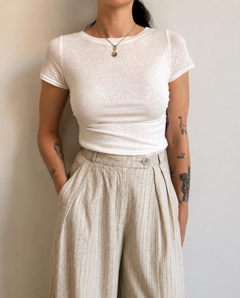 May include: A white short-sleeved crew neck t-shirt paired with high-waisted beige trousers featuring vertical pinstripes, a single button closure at the waistband, and deep front pleats. The trousers have a wide-leg silhouette and side pockets.