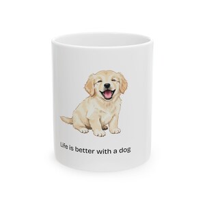 May include: White ceramic mug featuring a smiling golden retriever puppy illustration. The puppy has a light brown coat and a pink tongue. The text "Life is better with a dog" is printed below the illustration.