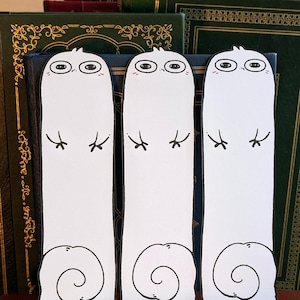 May include: Three white, whimsical bookmarks with cartoon ghost-like figures. Each bookmark features a large, oval-shaped head with eyes, eyelashes, and a small blush. The bottom of each bookmark has a spiral design.