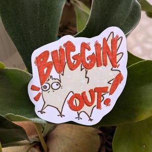 May include: A white sticker with the words "BUGGIN OUT!" in red, cartoon-style lettering. The sticker features a cartoon bug with big eyes and small legs. The sticker is set against a background of green leaves.