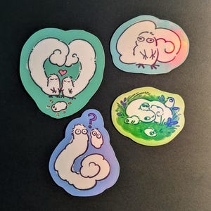 May include: Four whimsical stickers with cartoon cloud-like creatures. One sticker features two creatures with a heart, another shows a creature with a rainbow effect, and two more depict creatures in a grassy setting. The stickers have a white background.