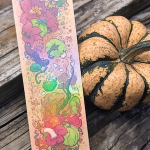May include: A rectangular bookmark featuring a colorful illustration of various green pumpkins, pink flowers, and small whimsical creatures on a gradient background transitioning from peach to yellow, displayed next to a speckled orange pumpkin.