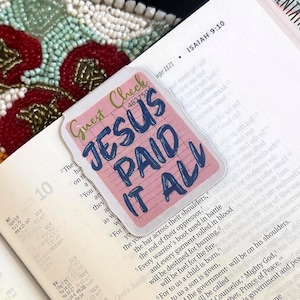 May include: An open Bible with a pink and blue sticker that reads "JESUS PAID IT ALL." The Bible is open to the book of Isaiah, with colorful tabs marking different books. A decorative beaded mat is in the background.