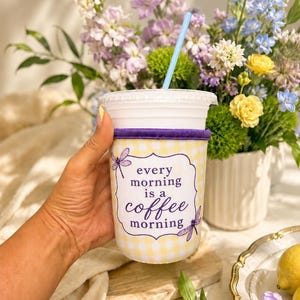 Puede incluir: Un vaso de plástico transparente con una pajita azul claro y una funda de tela de cuadros vichy amarillos y blancos. La funda tiene un borde morado y una etiqueta blanca con el texto "every morning is a coffee morning" y dos libélulas moradas.