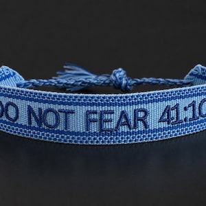Puede incluir: Pulsera azul tejida con el texto "DO NOT FEAR 41:10" en azul marino. La pulsera tiene un diseño tejido con borla y cierre de nudo. El fondo es oscuro.