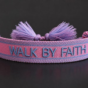 Op de afbeelding: Een roze en blauwe geweven armband met de geborduurde tekst "WALK BY FAITH". De armband heeft aan elk uiteinde een lichtpaarse kwast. De achtergrond is zwart.