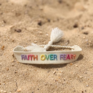 Puede incluir: Pulsera de tela color crema con las palabras bordadas "FAITH OVER FEAR" en colores arcoíris. La pulsera tiene una borla blanca y está sobre arena.