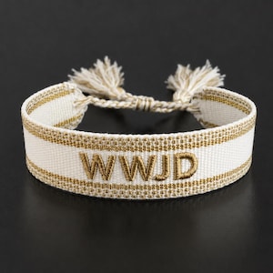 Pode incluir: Pulseira branca tecida com acabamento dourado e as letras bordadas "WWJD". A pulseira tem um fecho trançado com borlas bege. A pulseira é exibida em uma superfície preta.