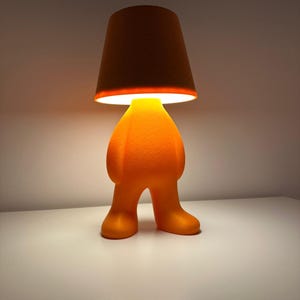 May include: Orange table lamp with a whimsical design. The lamp features a figure-shaped base with a conical lampshade. The lampshade is dark brown, and the base is a bright orange color. The lamp is on a white surface.