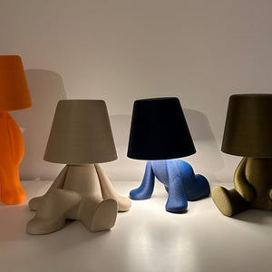 May include: Four whimsical table lamps in orange, beige, blue, and olive green. Each lamp features a shade atop a stylized human-like form in a seated or standing position. The lamps are arranged on a white surface.