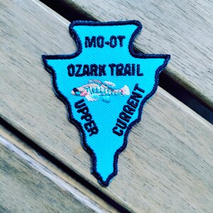 May include: An arrowhead-shaped embroidered patch in bright turquoise with a dark blue border. It features the black text MO-OT, OZARK TRAIL, UPPER, and CURRENT, along with a small, multi-colored embroidered fish graphic in the center.