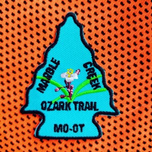 May include: An arrowhead-shaped embroidered patch in bright turquoise with a black border. It features a white and purple flower graphic with green leaves. The text reads MARBLE CREEK, OZARK TRAIL, and MO-OT in black, bold, capital letters.