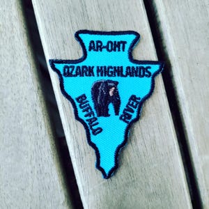May include: An arrowhead-shaped embroidered patch in bright cyan with a black border. It features the black text AR-OHT OZARK HIGHLANDS BUFFALO RIVER and a central black bear graphic. The design is centered on the patch with clear, bold lettering.