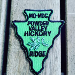 May include: A green and black embroidered patch with the text "MO-MDC POWDER VALLEY HICKORY RIDGE". The patch has a stylized arrowhead shape and features a light green embroidered leaf design.