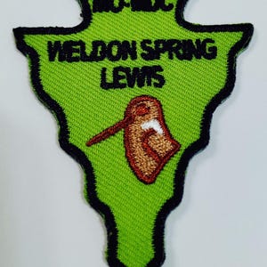 May include: A green embroidered patch with black trim in an arrowhead shape. The patch features the text "MO-MDC", "WELDON SPRING", and "LEWIS" in black. A brown and white graphic is in the center.