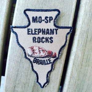 May include: An arrowhead-shaped embroidered patch with a tan background and dark blue border. The text reads MO-SP ELEPHANT ROCKS BRAILLE in dark blue thread. A small, detailed embroidered graphic of a rock formation is centered below the text.