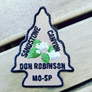 May include: An arrowhead-shaped embroidered patch with a tan background and dark blue border. It features green leaves and white flowers in the center, with the text SANDSTONE CANYON, DON ROBINSON, and MO-SP embroidered in dark blue lettering.