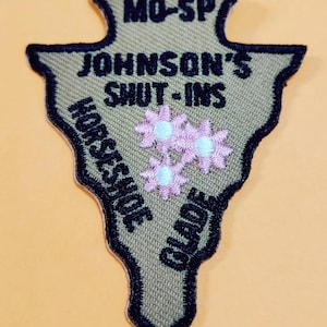 May include: Arrowhead-shaped patch with the text "MO-SP JOHNSON'S SHUT-INS HORSESHOE GLADE". The patch is olive green with a black border and features three light purple flower designs.