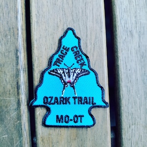May include: A turquoise arrowhead-shaped embroidered patch featuring a black butterfly graphic in the center. The text reads Trace Creek, Ozark Trail, and MO-OT in black embroidery. The patch has a black stitched border around the entire edge.