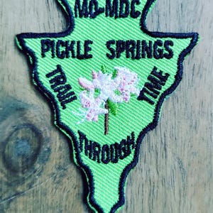 May include: An arrowhead-shaped embroidered patch in bright lime green with a black border. It features the black text MD-MDC, PICKLE SPRINGS, TRAIL TIME, and THROUGH, along with an embroidered illustration of a pink and white flower cluster.