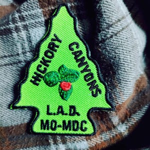 May include: A neon green embroidered patch in the shape of a stylized tree. The patch features the words "HICKORY CANYONS" and "L.A.D. MO-MDC" in black text. A small embroidered holly design with a red berry is in the center.