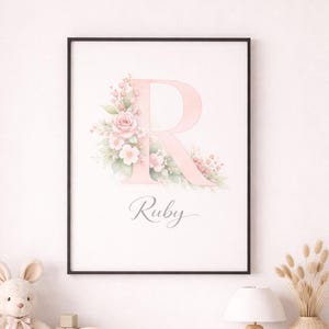 May include: A framed print featuring a large pink letter "R" adorned with floral accents and the name "Ruby" in cursive. The print is displayed above a white dresser with decorative items, including a stuffed bunny, rainbow, and wooden elephant lamp.