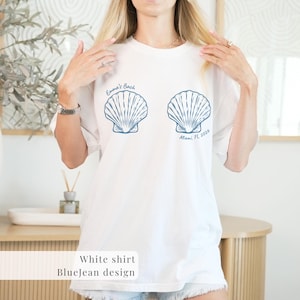 May include: White t-shirt with a blue seashell design. The shirt features two seashell illustrations and text that reads "Emma's Bach" and "Miami, FL 2026". The shirt is a casual, beach-themed design.