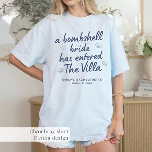 Op de afbeelding: Lichtblauw chambray shirt met de marineblauwe tekst "a bombshell bride has entered The Villa". Het shirt bevat ook "EMILY'S BACHELORETTE MIAMI, FL 2026". Het shirt heeft een denim design.