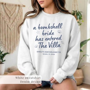 Op de afbeelding: Witte sweatshirt met marineblauwe tekst: "a bombshell bride has entered The Villa", met ster- en schelpgraphics. Daaronder staat "EMILY'S BACHELORETTE MIAMI, FL 2026". De onderkant van de afbeelding zegt "White sweatshirt Denim design".