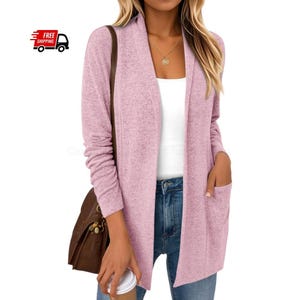 Könnte beinhalten: Ein hellrosa Cardigan mit offenem Schnitt, langen Ärmeln und zwei Vordertaschen. Der Cardigan ist aus einem weichen, strukturierten Stoff gefertigt. Das Model trägt ein weißes Oberteil und blaue Jeans. Eine braune Handtasche ist über der Schulter.