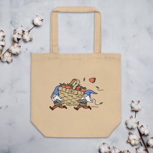 May include: Beige tote bag featuring an embroidered illustration of two gnomes carrying a basket overflowing with red strawberries. The gnomes wear blue hats and brown pants. A strawberry and green leaves are scattered around the basket.
