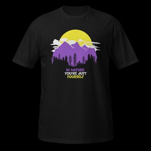 Puede incluir: Una camiseta negra de manga corta con un gráfico de montañas moradas, un sol amarillo y la silueta de un bosque negro. Debajo del paisaje, el texto dice: IN NATURE YOU'RE JUST YOURSELF en letras moradas, blancas y amarillas.