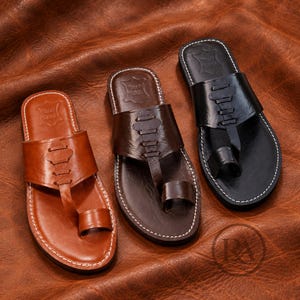 May include: Three leather sandals in tan, dark brown, and black. Each sandal features a wide strap across the top with decorative stitching, a toe loop, and a flat sole with white contrast stitching along the edge and a stamped logo on the heel.