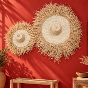 May include: Two circular wall decorations made of woven light-coloured straw with a raised centre and a wide, frayed, spiky fringe edge. The pieces are mounted on a solid red wall, featuring a natural texture and a sunburst-like appearance.