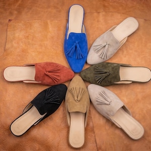 May include: A collection of seven slip-on mules arranged in a circular pattern. Each shoe features a pointed toe, a flat sole, and a decorative tassel on the upper. The colours include bright blue, light grey, olive green, beige, black, rust, and tan.
