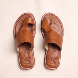 Peut inclure: Une paire de sandales en cuir marron clair avec une large bride à boucle d'orteil et une semelle intérieure profilée. Les bords des sandales sont finis avec des coutures blanches et un petit logo est embossé au niveau du talon.