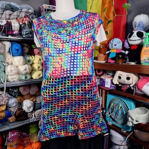 Puede incluir: Un top de crochet sin mangas y tejido abierto en un vibrante arcoíris de colores. El top se muestra en un maniquí y presenta un patrón de cuadrícula cuadrada. La paleta de colores incluye tonos de rojo, naranja, amarillo, verde, azul y morado.