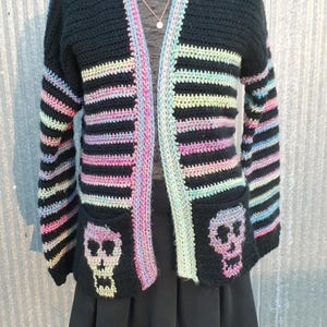 May include: Black crochet cardigan with colorful stripes and skull pockets. The cardigan is open in the front and has long sleeves.