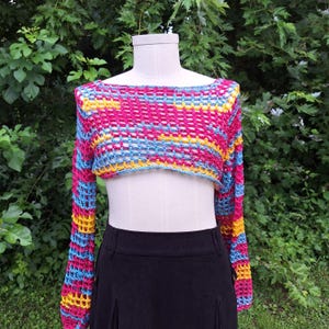 May include: A colorful, open-weave crochet top with long sleeves. The top features a vibrant mix of pink, blue, and yellow horizontal stripes. The garment is displayed on a mannequin, showcasing its cropped design.